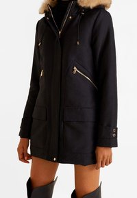 Zwarte parka met nepbont langs de capuchon, zakken met rits en gouden accenten. Voorzien van een ritssluiting aan de voorkant en verstelbare koordjes.