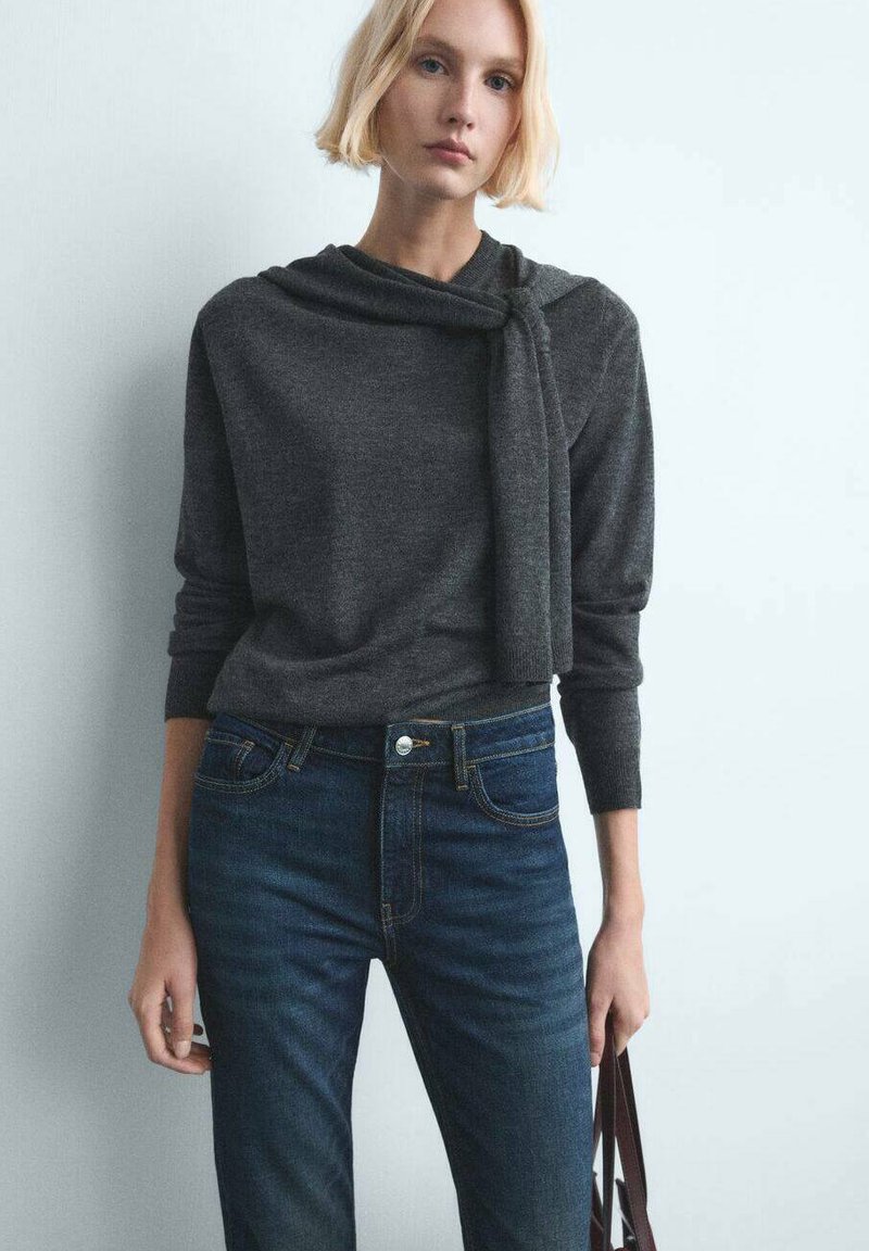 Mujer rubia que lleva un suéter de manga larga gris oscuro anudado en el hombro y jeans azules, sosteniendo una bolsa marrón a su lado.