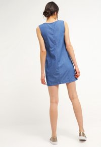 Džínové šaty s bezrukávovým designem, světle modré barvy, po kolena a se rovnými zadními švy. V kombinaci s metalickými pantoflemi.