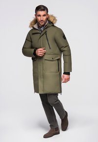 Mann trägt grüne lange Winterparka mit Fellkapuze, dunkle Hose und braune Wildlederstiefel, steht vor einem schlichten hellen Hintergrund.