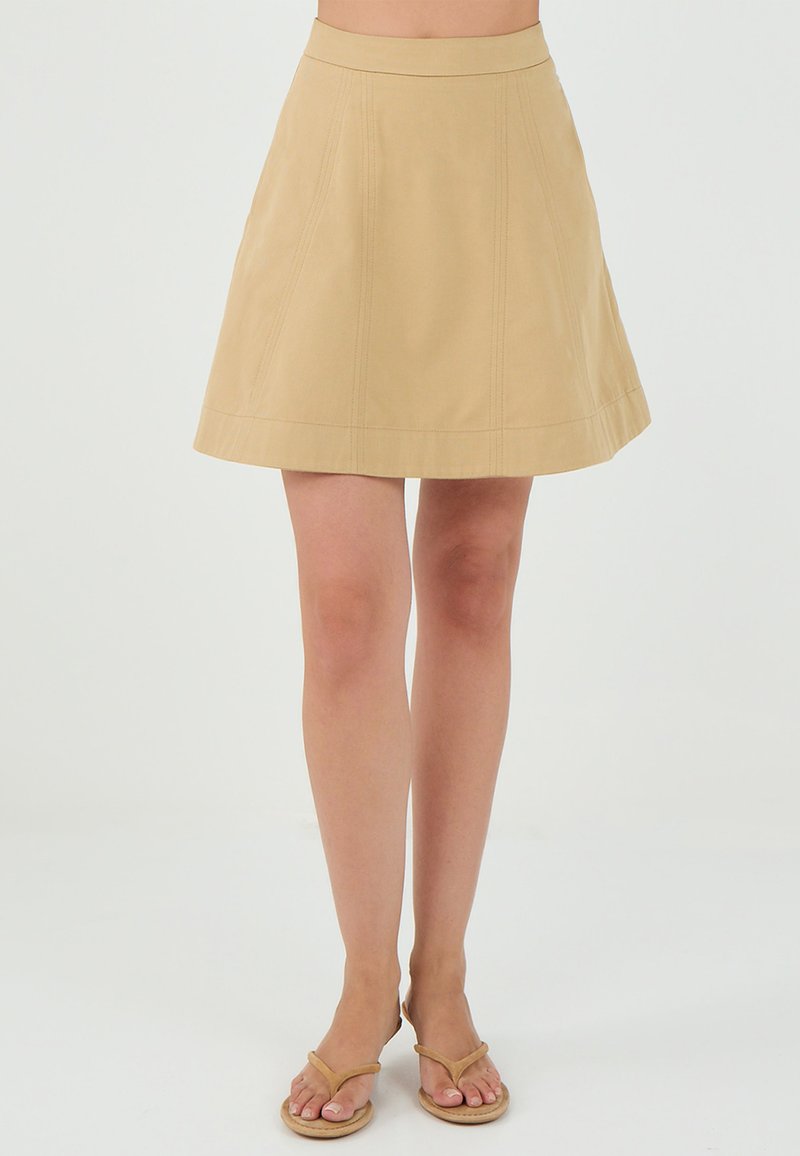 Jupe évasée beige avec une texture lisse, dotée de coutures horizontales et d'une taille ajustée. Une paire de sandales légères visible en bas.