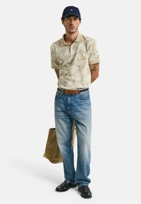 Beige kortærmet polo med tropemønster, lyseblå lige ben jeans, sorte sko, brun bælte og en mørk kasket; holder en beige taske.