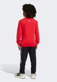 Red sweatshirt with long sleeves, ribbed cuffs, and a logo at the neck, paired with black sweatpants featuring white side stripes and sneakers.