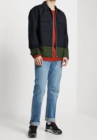 Black and green zip-up jacket with two front pockets, layered over a red shirt, paired with blue jeans and black athletic shoes.