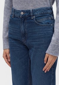 Gros plan d'une personne portant un jean bleu taille haute et un pull tricoté gris avec les mains détendues le long du corps.