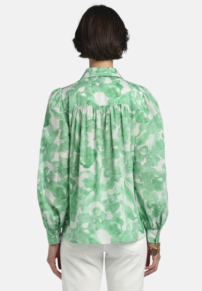 Sieviete, kas valkā zaļu un baltu ziedu raksta blūzi ar gariem pūstiem piedurknēm un baltas bikses, redzama no mugurpuses uz vienkrāsaina fona.