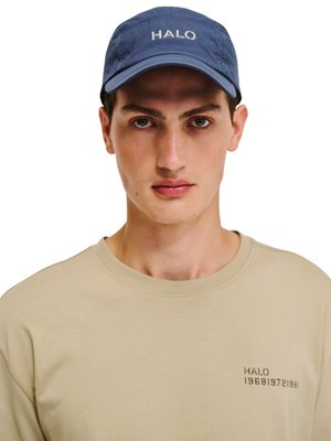 Młody mężczyzna noszący niebieską czapkę z napisem „HALO” oraz beżową koszulkę z małym napisem „HALO” i cyframi na lewej piersi, zwrócony przodem.