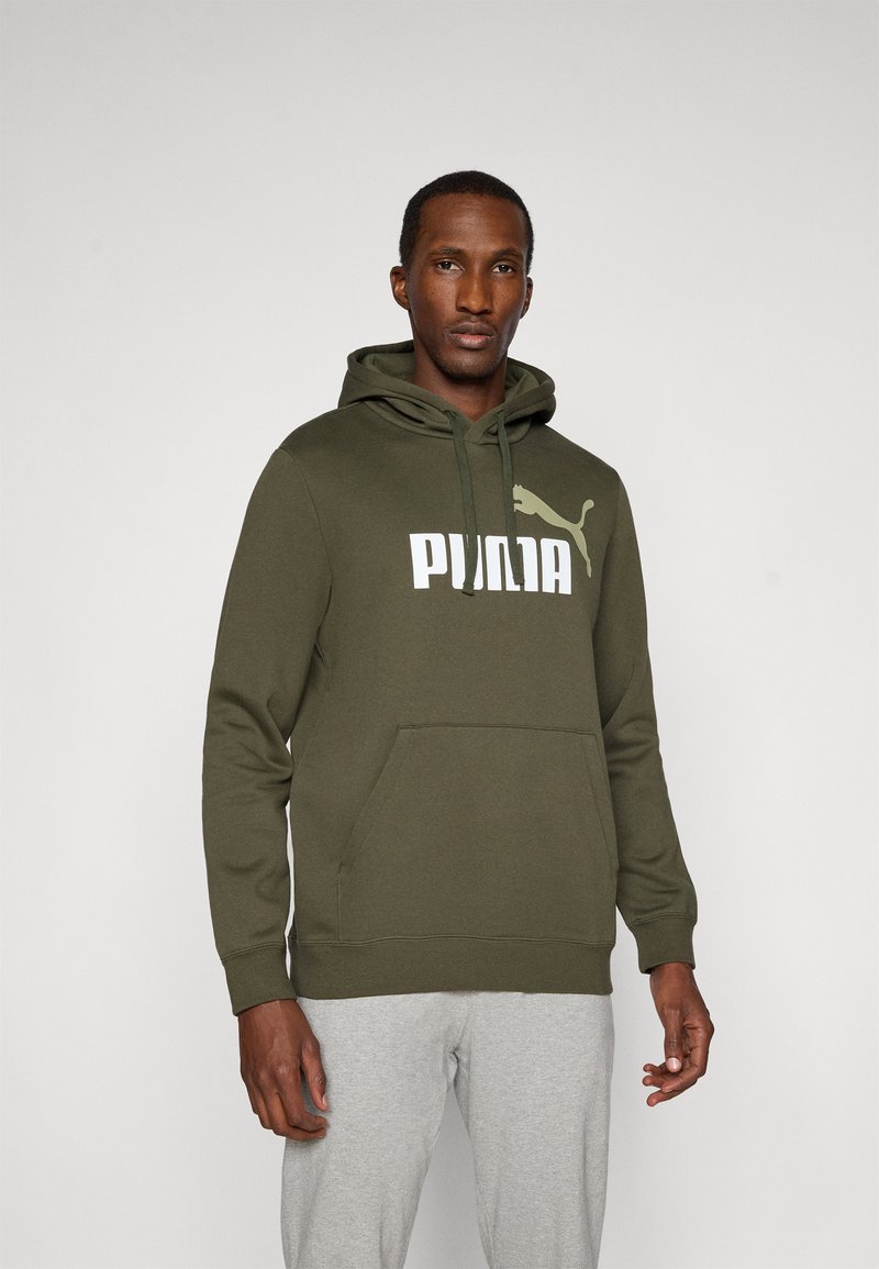 Mand iført en olivengrøn Puma hoodie med frontlomme og lysegrå svedbukser, stående foran en ensfarvet lys baggrund.
