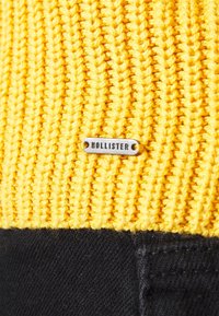 Tejido de punto amarillo con canalé vertical; presenta una etiqueta plateada grabada con "HOLLISTER" sujeta a una sección negra debajo.