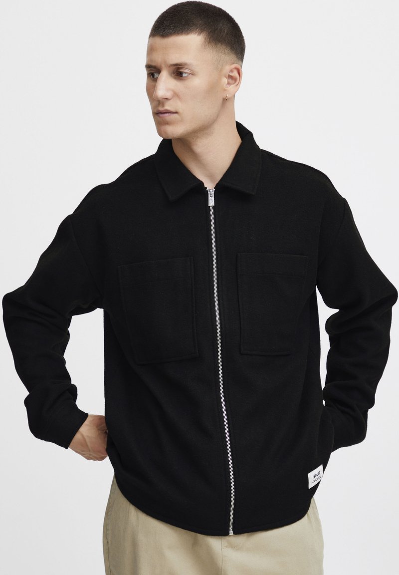 Black zip-up jacket made of soft fabric; features two front pockets and a collar. Smooth texture with a relaxed fit. Tag visible at the hem.