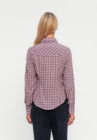 Chemise à carreaux violette et blanche, avec un col pointu, un ourlet arrondi et des poignets à boutons. La vue de dos montre le détail de l'empiècement et la coupe.