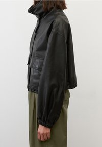 Donna che indossa una giacca di pelle nera corta con polsini elasticizzati e pantaloni verde oliva, mostrata di lato su uno sfondo neutro.