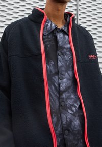 Černá fleecová bunda s červenými zipovými detaily přes černou a šedou marbled košili s knoflíky. Zřetelná textura a kontrastní vrstvy.