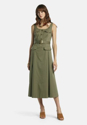 Femeie purtând o rochie midi fără mâneci, verde măsliniu, cu centură și buzunare cu clapetă, mule maro cu toc, colier și brățară de chihlimbar, stând pe fundal alb.