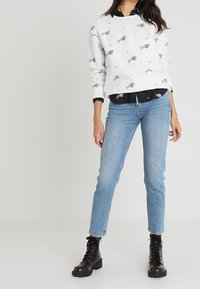 Sudadera blanca con estampados de peces y flores, superpuesta a una camisa negra con cuello, combinada con jeans de mezclilla azul claro y botas negras con cordones.