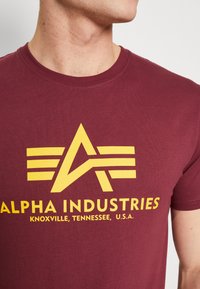 Rūsganas t-krekls, kas izgatavots no mīksta auduma, ir ar lielu dzeltenu ģeometrisku logo, ar svītrām un tekstu "ALPHA INDUSTRIES" zem tā.