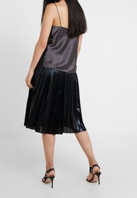 Camisola nera di satin con spalline sottili abbinata a una gonna nera plissettata, caratterizzata da una texture lucida e un design a metà lunghezza. Sandali con tacco completano l'outfit.