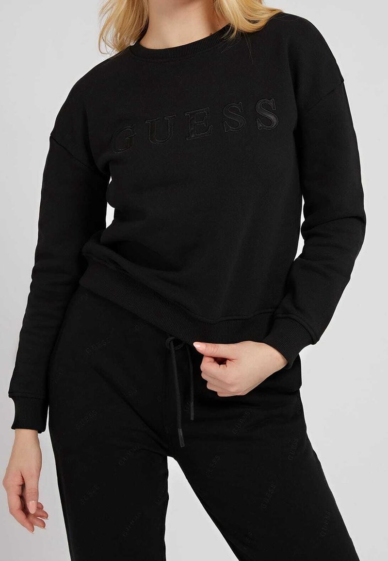 Sweatshirt noir avec logo "GUESS" en relief. Comprend un col rond, des manches longues et une coupe décontractée. Associé à un pantalon noir assorti.