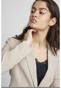 Blazer beige con rever a punta, tessuto texture e tre bottoni chiari sul maniche. Indossato su un top in pizzo nero.