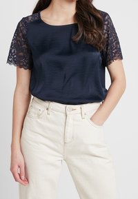 Mörkblå satinblus med korta spetsärmar, med rund halsringning och avslappnad passform, kombinerad med ljusa högmidjade byxor.