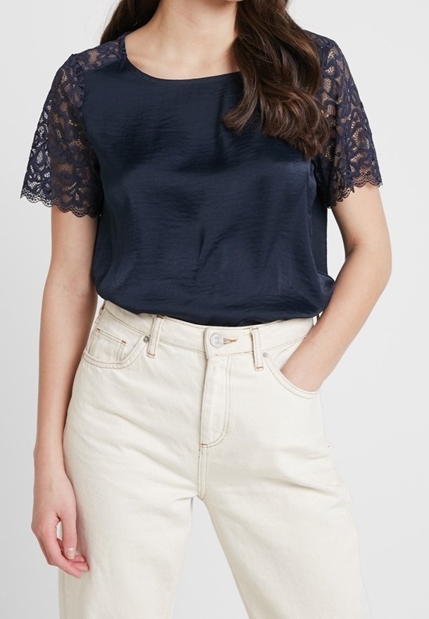 Mörkblå satinblus med korta spetsärmar, med rund halsringning och avslappnad passform, kombinerad med ljusa högmidjade byxor.