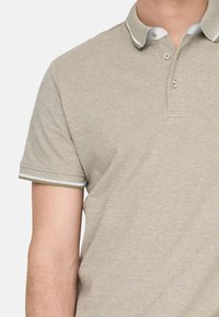 Beige Poloshirt mit strukturierter Oberfläche. Hat einen Kragen mit kontrastierenden weißen und grünen Streifen sowie eine kurze Knopfleiste mit zwei Knöpfen. Kurze Ärmel.