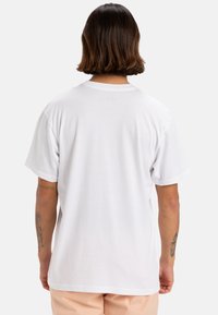 Camiseta blanca de algodón con mangas cortas, diseño liso, cuello redondo y un corte relajado. Incluye tatuajes visibles en los brazos.