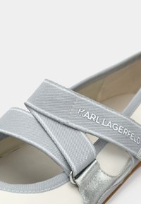 Γκρι υφασμάτινο παπούτσι με σχέδιο σταυρωτού λουριού, που διαθέτει μεταλλική λεπτομέρεια και μπεζ εσωτερικό. Το όνομα της μάρκας "Karl Lagerfeld" είναι ορατό.