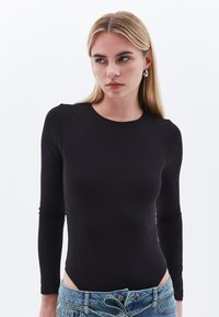 Schwarzer langärmeliger Bodysuit mit rundem Ausschnitt und weicher Textur. Besitzt seitliche Ausschnitte. Kombiniert mit blauen Denim-Shorts.