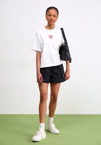 White oversized t-shirt with red heart emblem, black athletic shorts, white sneakers, and black bag. Green floor background.