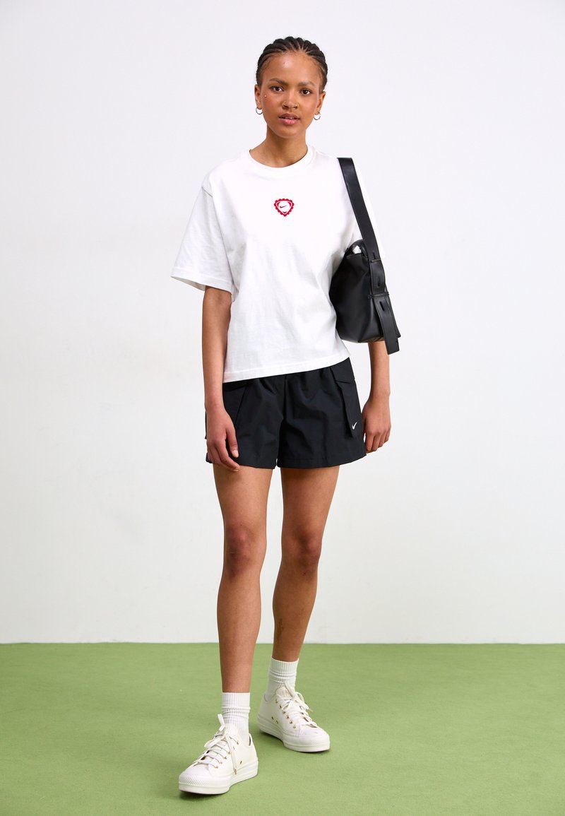 Weißes oversized T-Shirt mit rotem Herzsymbol, schwarze Sportshorts, weiße Sneaker und schwarze Tasche. Grüner Boden als Hintergrund.