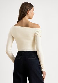 Hosszú barna hajú nő, váll nélküli, krémszínű hosszú ujjú felsőt és sötét, magasderekú nadrágot viselve, oldalra néz egy egyszerű háttér előtt.