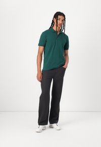 Dunkelgrünes Polohemd mit Kragen und einem Halb-Reißverschluss, kombiniert mit locker sitzenden, dunkelgrauen Hosen und weißen Sneakers.