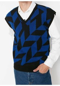 Uomo che indossa un gilet senza maniche a motivi geometrici blu e neri sopra una camicia bianca con bottoni e jeans blu, con una mano in tasca.
