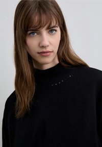 Svart tröja med hög hals och ett delikat mönster av små utskärningar längs halsringningen, tillverkad av ett mjukt, texturerat tyg.