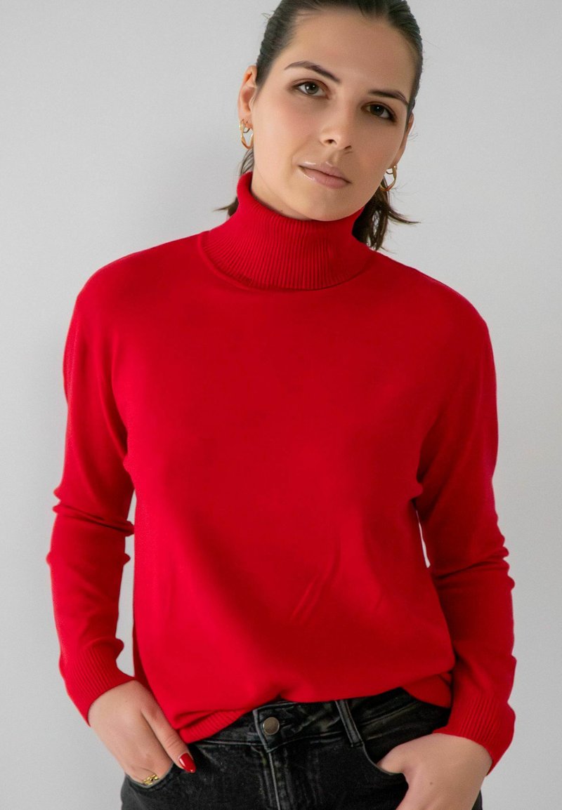 Pull à col roulé rouge en maille douce, avec un col et des poignets côtelés, porté sur un jean sombre, présentant une texture lisse et une coupe décontractée.