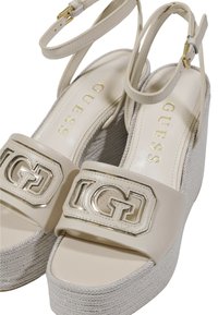 Sandali a zeppa beige con fibbia luccicante a logo, tomaia in pelle, piattaforma con texture intrecciata e cinturini alla caviglia. Accenti dorati arricchiscono il design.