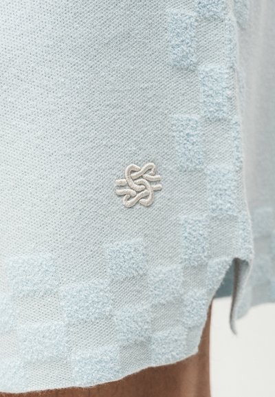 Υφασμα πλεκτό σε ανοιχτό μπλε χρώμα με υφή, διακριτικό καρό μοτίβο και μικρό λευκό κεντημένο λογότυπο κόμπου κοντά στην άκρη.