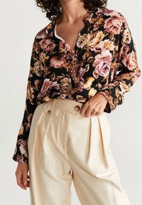 Blusa floreale nera, con rose rosa e gialle, dotata di bottoni e maniche lunghe. Pantaloni plissettati crema con dettagli in bottoni.