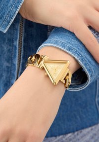 Gold triangle watch with a shiny finish, featuring a chunky link bracelet. The design combines geometric shapes and smooth textures.