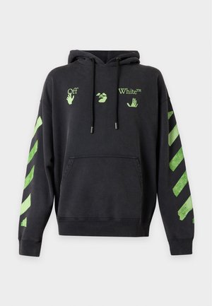 Homme portant un sweat à capuche noir avec des manches à rayures vertes et un texte imprimé, associé à un pantalon sombre à motifs et des baskets noires.