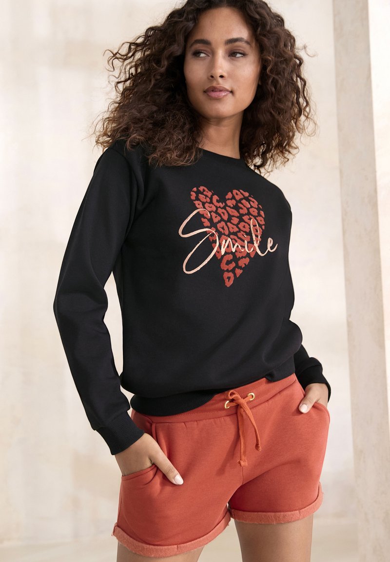 Mustapaita, jossa on leopardikuvioinen sydän ja "Hymyile" -teksti, yhdistettynä oransseihin nyörishortseihin, joissa on käännetyt lahkeet ja taskut.