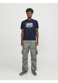 Camiseta de color azul marino con un estampado gráfico en rojo y blanco. Pantalones cargo grises con bolsillos laterales. Zapatillas grises con una superficie texturizada.