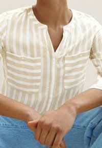 Personne portant une chemise rayée verticale beige et blanche avec deux poches avant, assise les mains entrelacées sur un jean bleu.