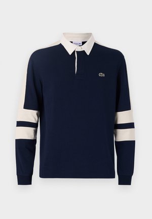 Polo à manches longues marine avec un col blanc. Présente des accents rayés crème et marine sur les manches. Logo Lacoste sur la poitrine gauche.
