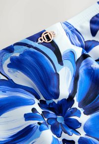 Kwiatowa tkanina z niebieskimi i białymi płatkami o gładkiej fakturze. W pobliżu szwu widoczny jest detal z różowego złota.