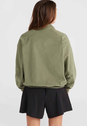 Olivnozeleni flisanj jacket z visokim ovratnikom in raglan rokavi, v kombinaciji s črnimi kratkimi hlačami. Mehka tekstura in sproščen dizajn.