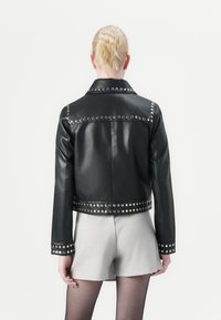 Svart fusk lærjakke med sølvnitter langs skuldrene og kanten, med krage og en kort design, kombinert med lyse shorts.