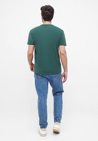 Donkergroene T-shirt met korte mouwen, relaxte pasvorm, gecombineerd met lichtblauwe denim jeans en witte sneakers met beige accenten, gezien van achteren.