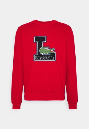Rød sweatshirt med lange ærmer, ribbede manchetter og rund hals. Har et stort broderet "L" og krokodillelogo med sorte detaljer.